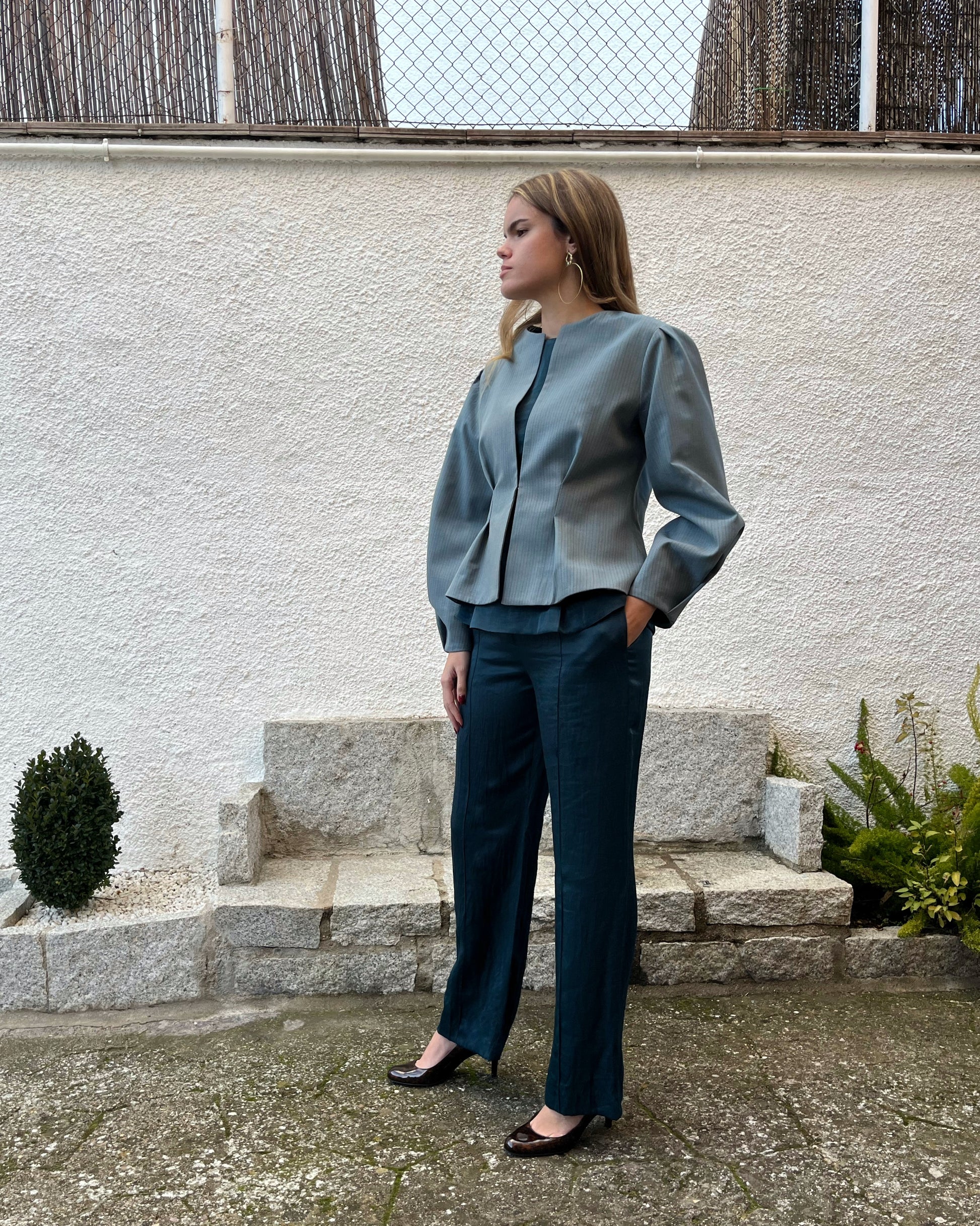Vista lateral de chaqueta azul elegante de mujer con rayas a tono y mangas abullonadas; modelo posando en jardín.