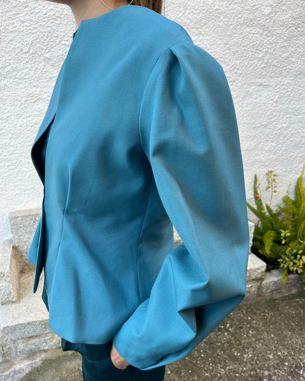 Vista lateral de chaqueta azul turquesa de mujer con mangas abullonadas y cierre oculto; modelo en jardín.