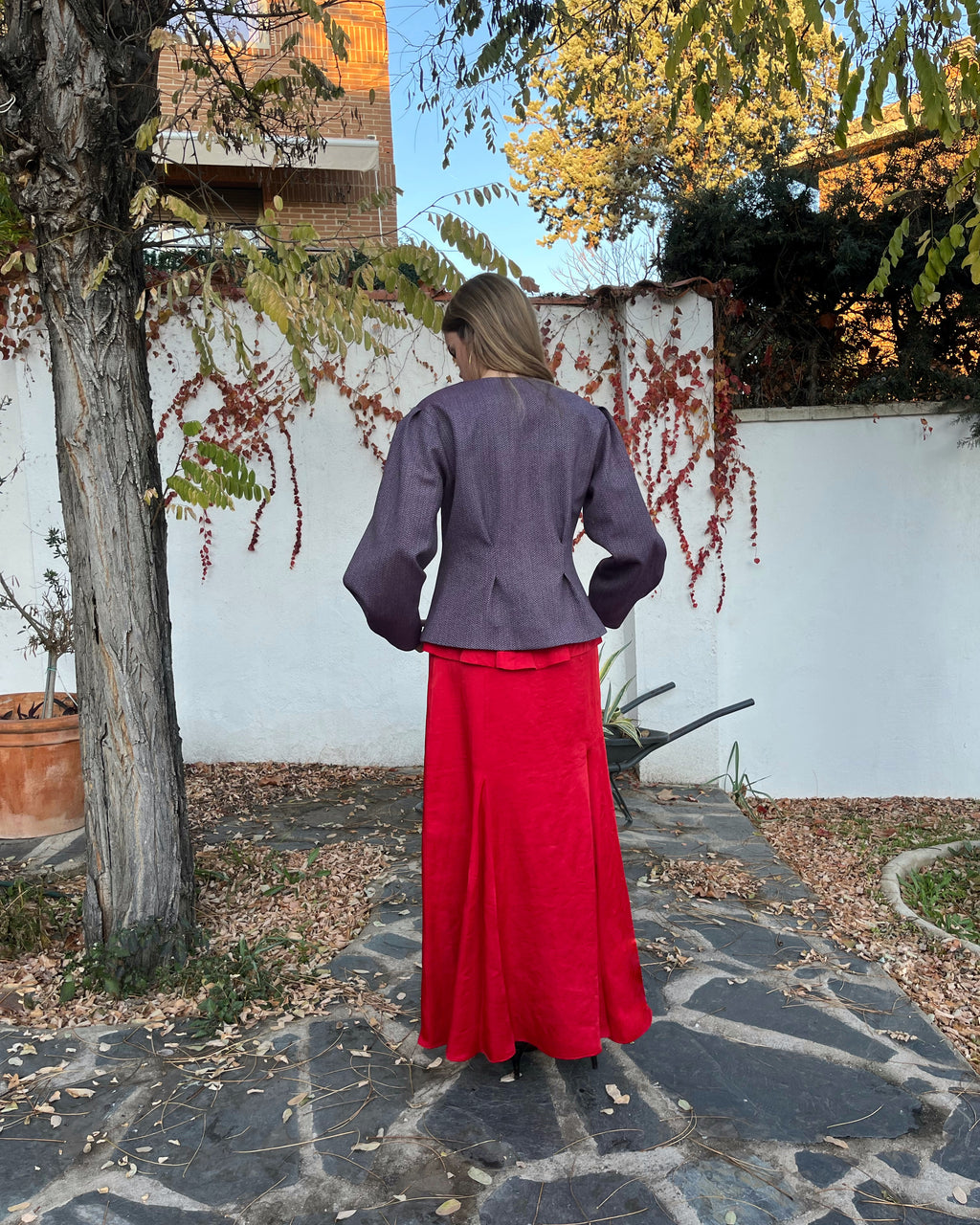 Chaqueta morada de mujer con tejido de espiga y pliegues entallados; vista trasera en jardín, estilo elegante.