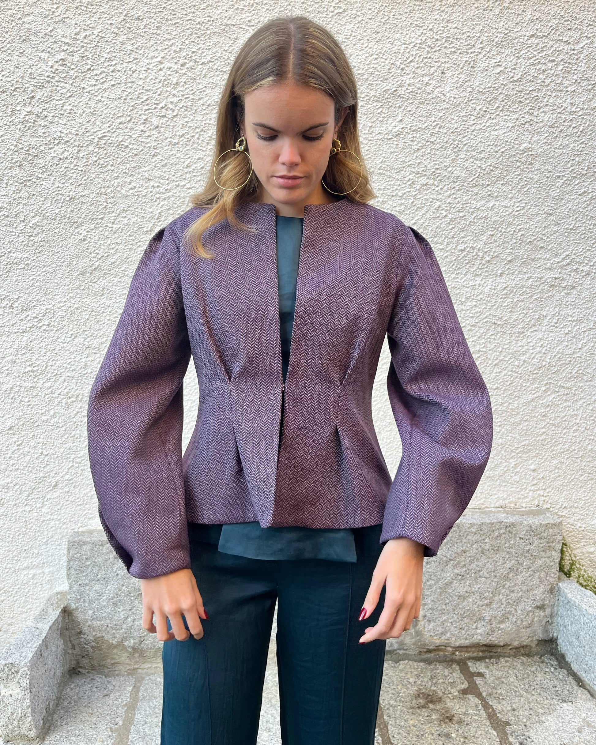 Chaqueta morada de mujer con textura de espiga y diseño elegante entallado; vista frontal con modelo en jardín, colección Historia de un sastre.