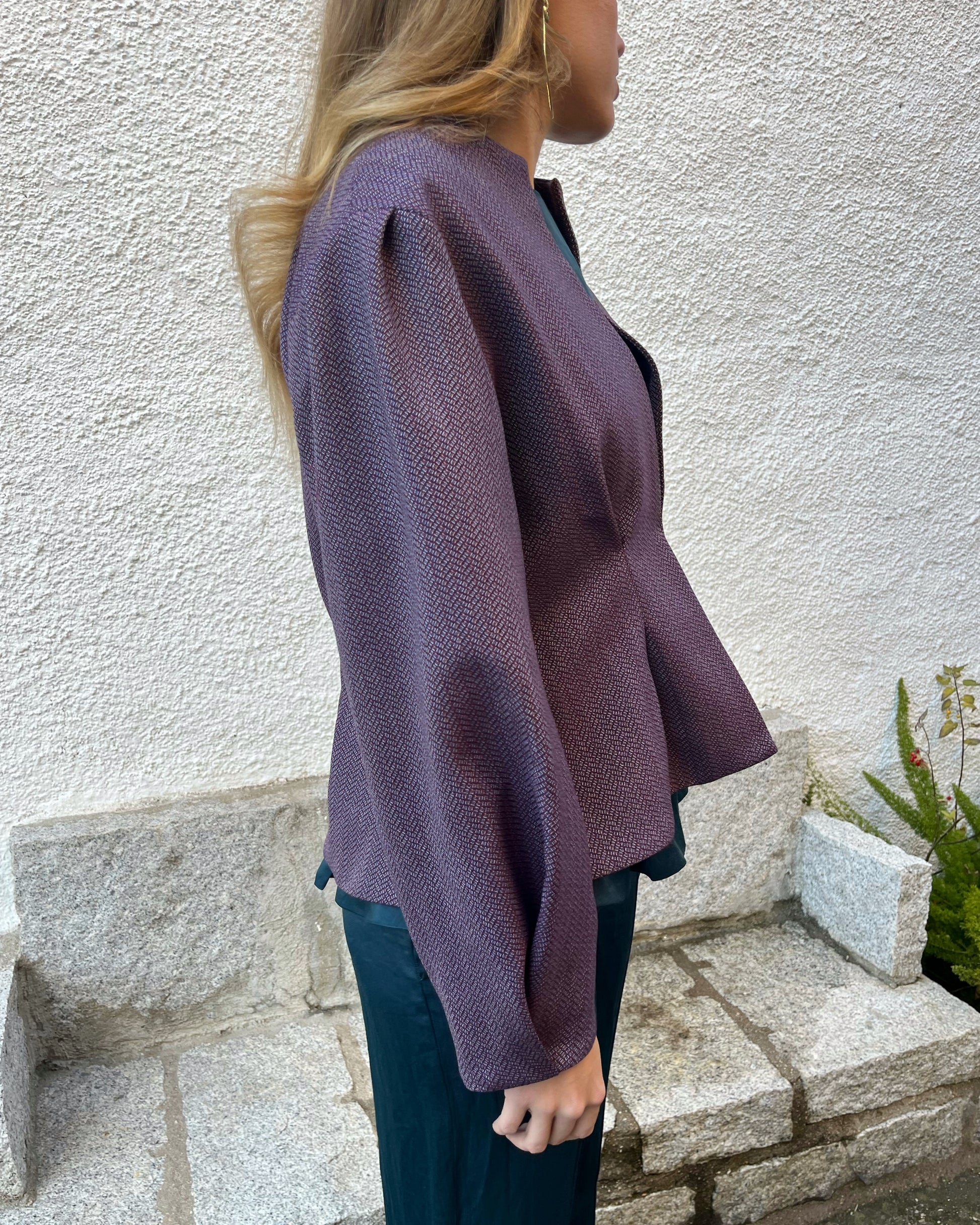 Vista lateral de chaqueta morada elegante con textura de espiga, mangas abullonadas y cierre oculto; modelo en jardín.