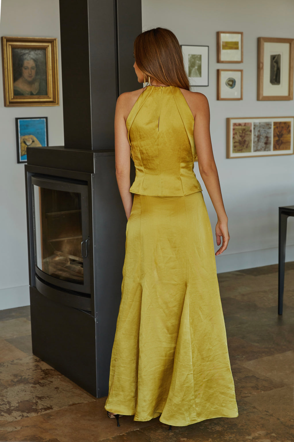 Vista trasera de top halter color aceite amarillo verde dorado elegante de mujer con escote lágrima, botón forrado y espalda entallada; modelo posando en jardín.