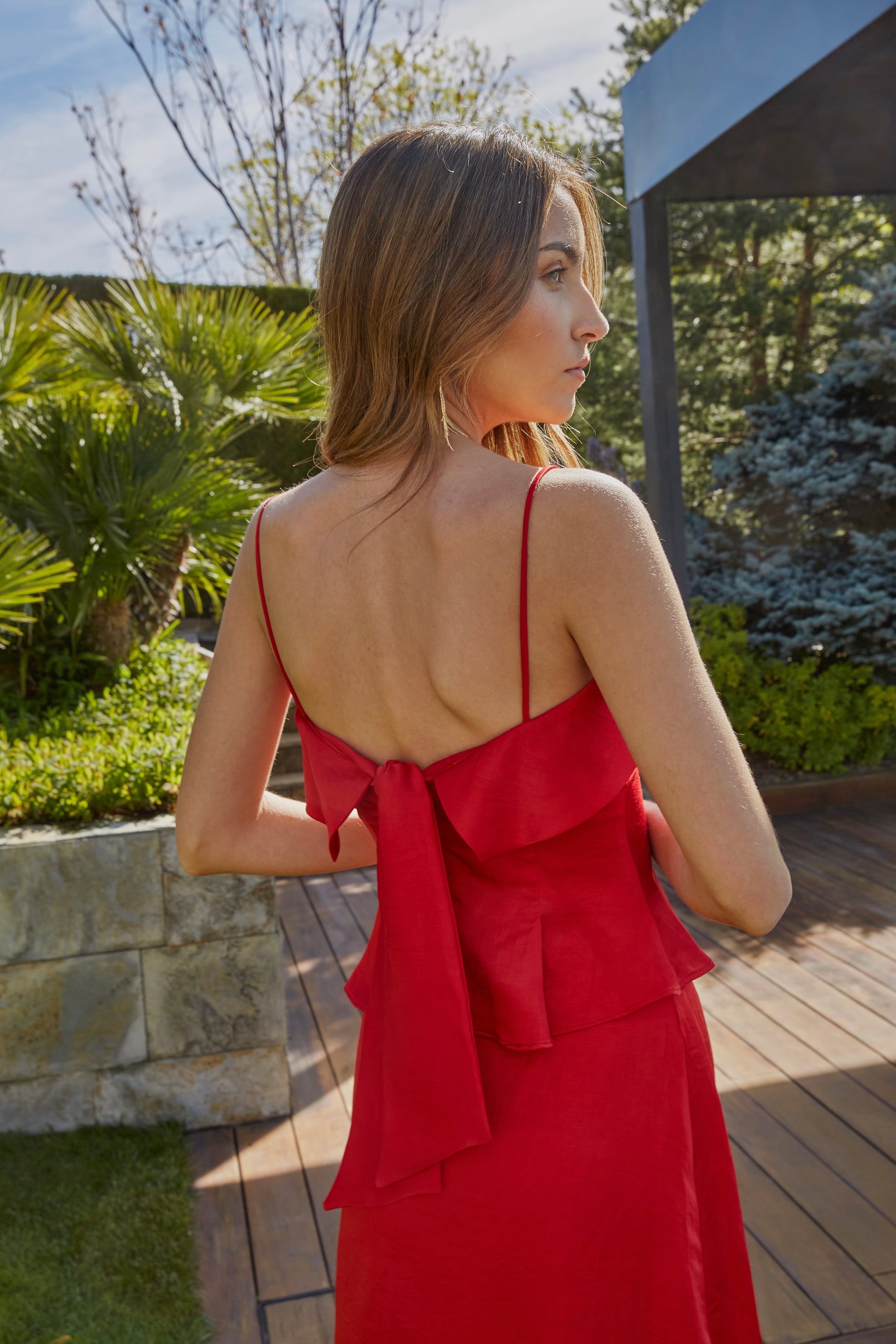 Vista trasera de top rojo elegante de mujer con tirantes finos y lazo en la espalda; diseño ideal para eventos y bodas en jardín.